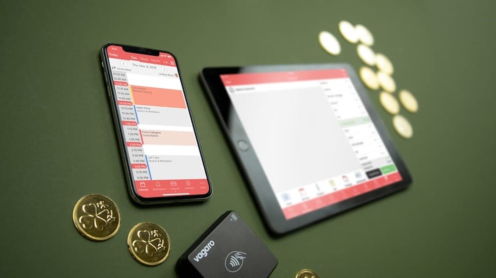 A phone, tablet, and Vagaro credit card reader sits on a green surface with gold coins around the pieces of tech.