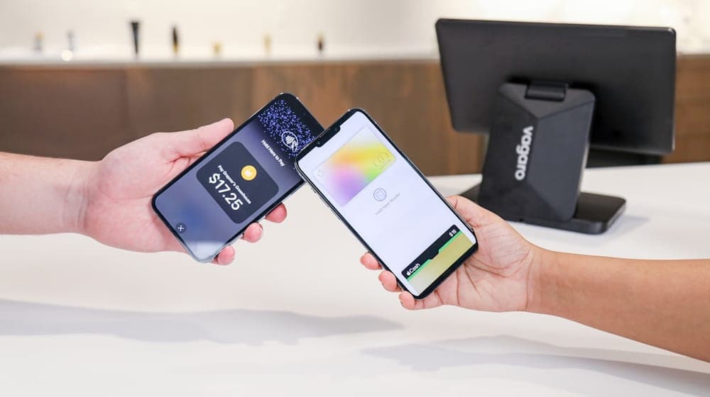 At the front desk of a salon, a customer uses their phone to pay a business owner for their service. The Vagaro PayPro hardware sits behind the transaction at a warm and well lit salon.