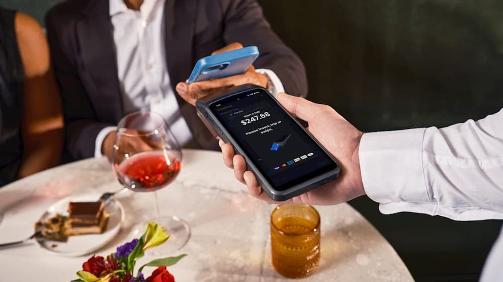 A close-up shows a fine dining guest tapping their smartphone to a SpotOn handheld POS terminal displaying a $247.68 checkout screen, with red wine and flowers on the marble table. SpotOn's elegant tableside payment experience is built for the demands of upscale hospitality. Learn more at spoton.com